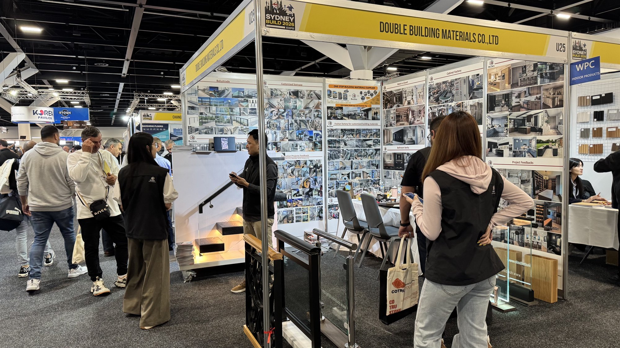 The Double Building Materials stand U25 at Sydney Build 2026, with a yellow header banner, project photo walls and glass and cast-iron railing samples, as visitors browse