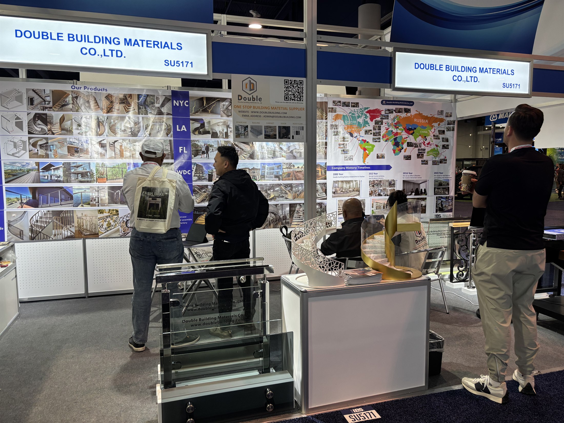 The Double Building Materials booth SU5171 at IBS 2025 in Las Vegas, with a world-project map wall, a gold spiral staircase model and a glass railing model, as visitors browse