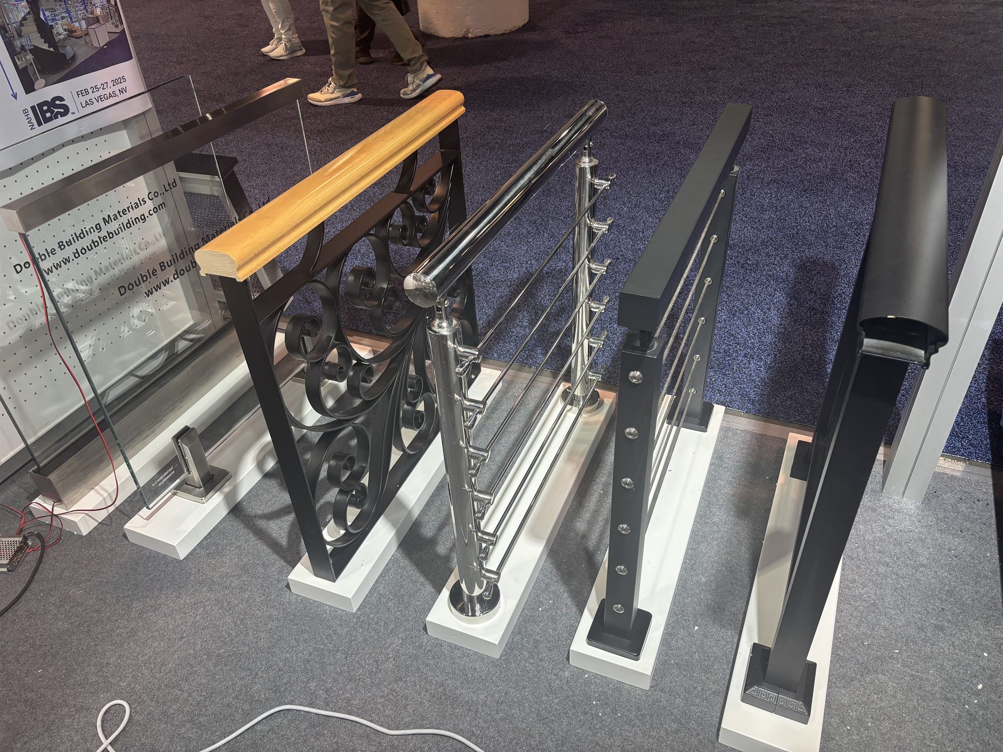 A row of railing post samples on the floor at IBS 2025, from an ornate cast-iron baluster with a wood handrail to stainless cable and powder-coated steel posts