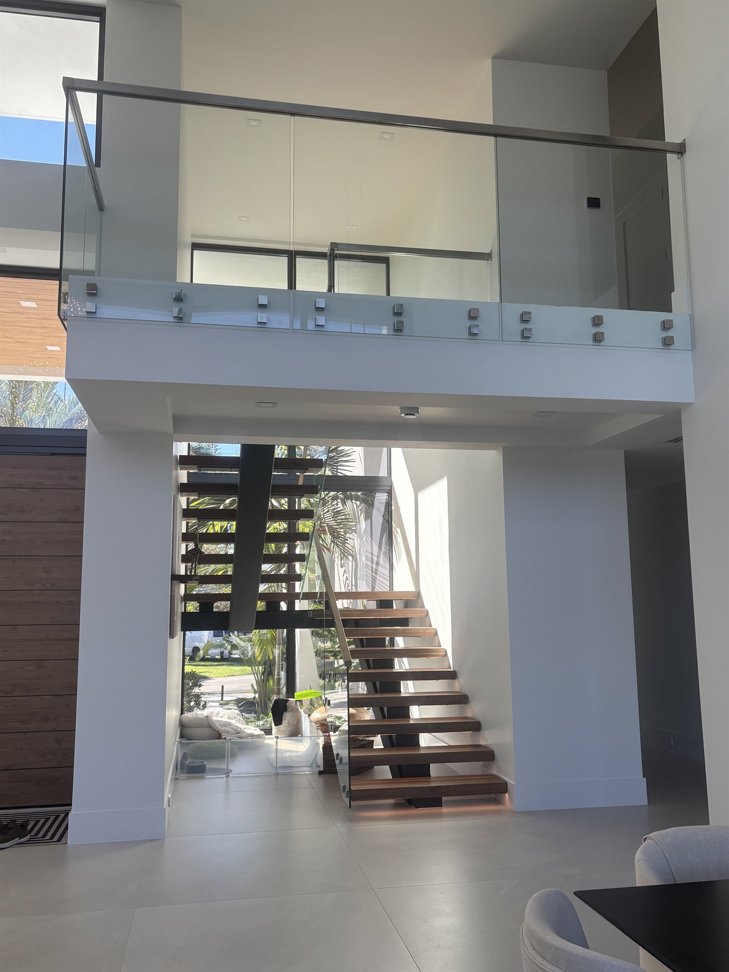 A double-height entry with a frameless glass balustrade on the upper landing and the floating wood staircase beyond, in a Fort Lauderdale home