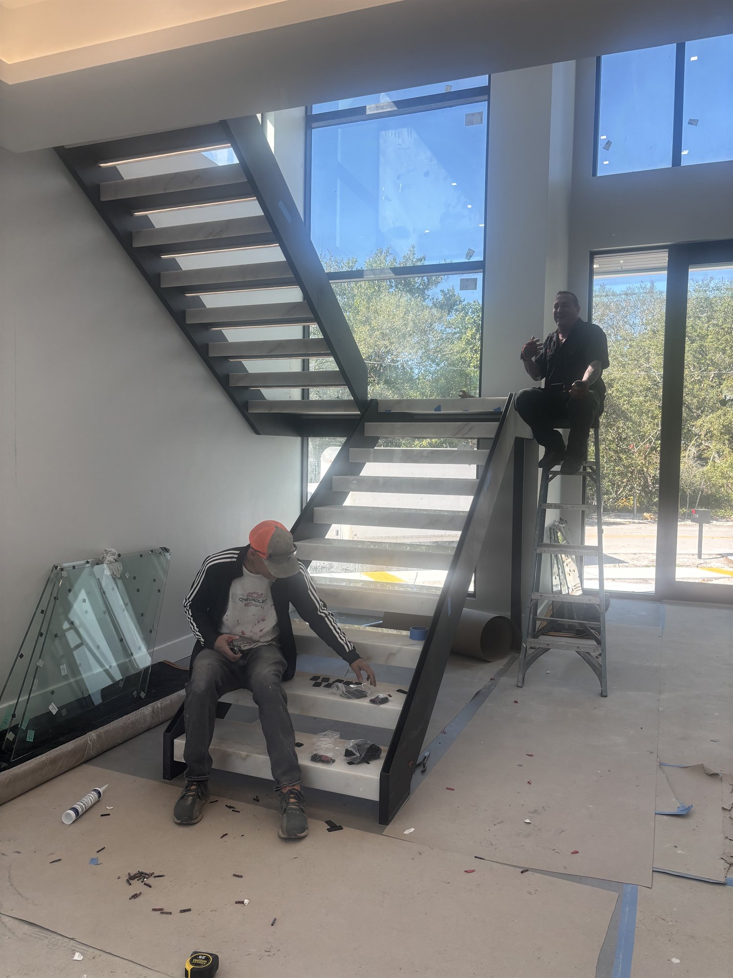 The floating walnut and black-steel staircase part way through fitting on site in Fort Lauderdale, with glass panels staged on the floor