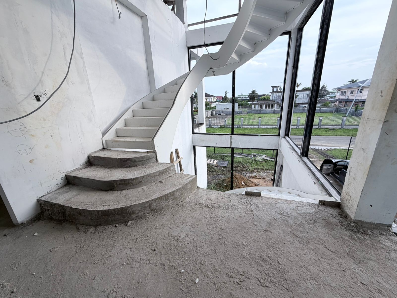 Curved staircase rising along a white wall beside a full-height glass facade during construction of a private home in Suriname