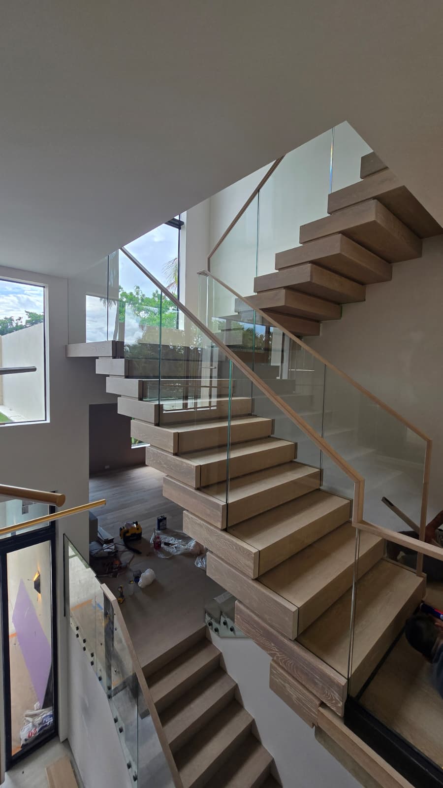 White-oak floating treads cantilevering past a frameless glass balustrade with an oak top cap, mid build, in a Florida home
