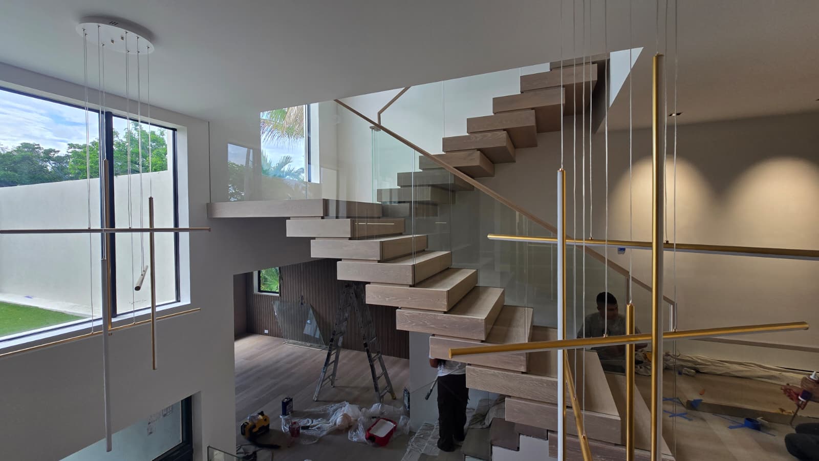 White-oak floating staircase with frameless glass and a linear pendant beside tall windows, mid fit-out in a Florida new-build home